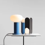 Mała lampa stołowa Flo Sky z białym kloszem o zaokrąglonym kształcie, umieszczona na niebieskiej podstawie. Obok znajduje się złożona dekoracja w odcieniach czerni i srebra. Lampa jest podłączona do zasilania, a całość stoi na białym, marmurowym blacie.