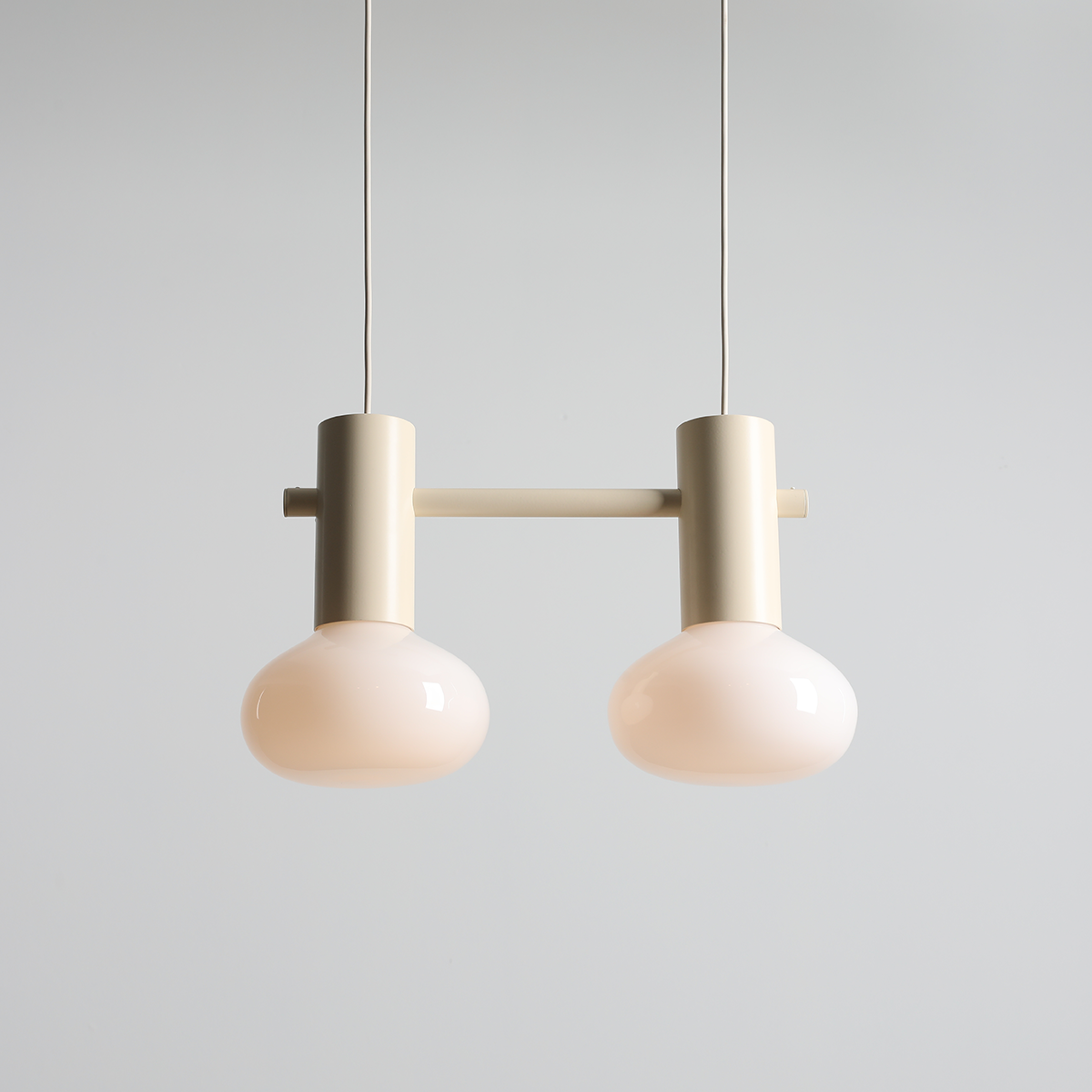Lampa wisząca Flo Cream z dwoma kloszami E14, w kolorze kremowym. Klosze mają zaokrąglony kształt, a całość podwieszona jest na cienkich linkach. Elementy metalowe w neutralnym odcieniu, minimalistyczny design. Tło jasne, proste.