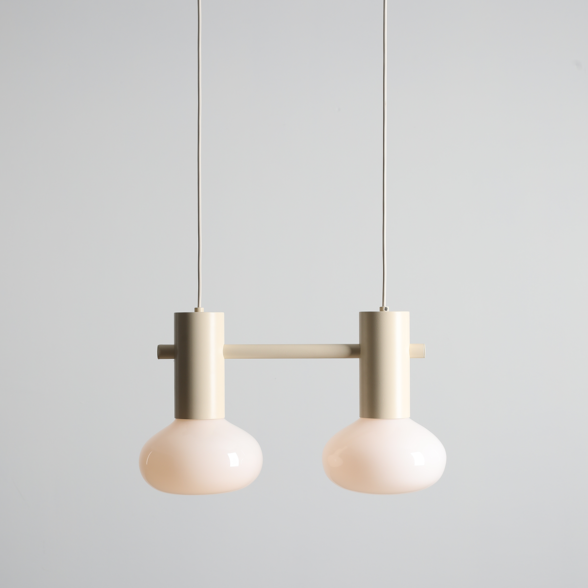 Lampa wisząca Flo Cream z dwoma kloszami z przezroczystego szkła w odcieniu jasnego różu, zawieszona na cienkich przewodach. Metalowa konstrukcja w kolorze kremowym, prosta forma z poziomym uchwytem. Tło jasne, minimalistyczne.