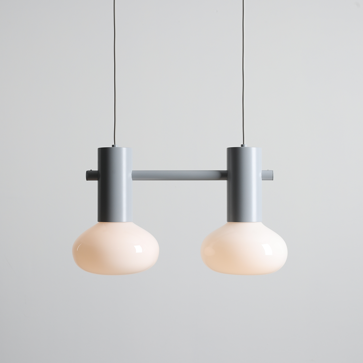 Lampa wisząca Flo Grey z dwoma kloszami w kolorze mlecznym, zawieszona na metalowym, szarym stelażu. Klosze mają zaokrąglony kształt, a lampa wisi na cienkim przewodzie w neutralnym otoczeniu.