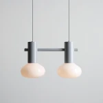 Lampa wisząca Flo Grey z dwoma kloszami w kolorze mlecznym, zawieszona na metalowym, szarym stelażu. Klosze mają zaokrąglony kształt, a lampa wisi na cienkim przewodzie w neutralnym otoczeniu.