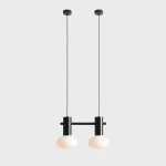 Lampa wisząca Flo Moss z dwoma oprawami E14, z ciemnozielonym metalowym stelażem i mlecznymi, okrągłymi kloszami. Ułożona w pionie, na długich przewodach, w minimalistycznym stylu, na jednolitym tle.