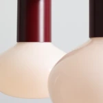 Lampa wisząca Flo Ruby z dwoma kloszami w kolorze jasnego różu, zawieszona na czerwonym metalowym mocowaniu. Klosze mają zaokrąglony kształt, tworząc nowoczesny design. Produkt umieszczony na jednolitym tle.