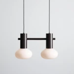 Lampa wisząca Flo Mokka z dwoma oprawami E14, o matowych, okrągłych kloszach w kolorze białym i metalowej konstrukcji w odcieniu ciemnego brązu. Zawieszona na cienkim, czarnym przewodzie na jednolitym tle.