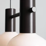 Lampa wisząca Flo Mokka z dwoma kloszami E14, w kolorze czarnym matowym. Klosze mają okrągły kształt, wykonane z białego szkła. Produkt zawieszony na cienkim, czarnym kablu, z widocznym elementem mocującym. Tło w jasnoszarym kolorze.