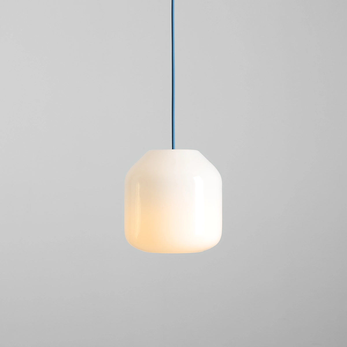 Lampa wisząca Flask Sapphire D18 z białym, zaokrąglonym kloszem i niebieskim przewodem. Klosz emituje ciepłe światło, zawieszony w centralnej pozycji na szarym tle. Idealna do nowoczesnych wnętrz.