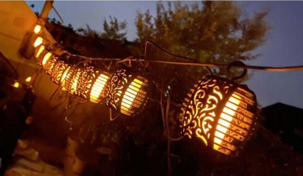 Girlanda ogrodowa solarna Flami z 20 LED, długość 3,5 m, czarna, zawieszona w nocy. Oświetlona lampionami w kształcie kul z dekoracyjnymi wzorami, emitującymi ciepłe, pomarańczowe światło, otoczona delikatnym tłem zieleni.