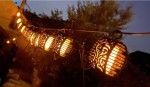 Girlanda ogrodowa solarna Flami z 20 LED, długość 3,5 m, czarna, zawieszona w nocy. Oświetlona lampionami w kształcie kul z dekoracyjnymi wzorami, emitującymi ciepłe, pomarańczowe światło, otoczona delikatnym tłem zieleni.