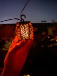 Girlanda ogrodowa solarna Flami z 20 diodami LED, czarna, zawieszona na sznurku. W kształcie lampionu z dekoracyjnym wzorem, emitująca ciepłe światło. Trzymana w dłoni, w tle widoczny zmierzch.