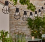 Girlanda ogrodowa solarna Lofti z 20 LED, czarna, zawieszona na lince. Klosze w kształcie żarówek, osłonięte metalową siatką, rozmieszczone równomiernie. Tło z zielonymi roślinami, stonowane i naturalne. Długość girlandy wynosi 4 m.