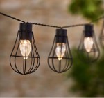 Girlanda ogrodowa solarna Lofti z 20 diodami LED, czarna, z metalowymi kloszami w kształcie kropli. Ujęcie zbliżone, ukazujące kilka lamp wiszących na czarnym przewodzie, w tle rozmyta zielona roślinność.
