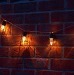 Girlanda ogrodowa solarna Lofti 20xLED, czarna, zawieszona na tle ceglanej ściany. Widoczne ciepłe światło emitowane przez żarówki w metalowych kloszach, rozmieszczone w regularnych odstępach na przewodzie. Długość girlandy wynosi 4 metry.