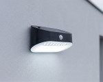 Czarny kinkiet elewacyjny Uran LED z czujnikiem ruchu i zmierzchu, zamontowany na jasnej ścianie. Posiada prostokątny kształt, z przezroczystą osłoną na diody LED, emitującą białe światło. Widoczny czujnik z przodu.