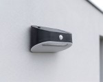 Czarny kinkiet elewacyjny Uran LED z czujnikiem ruchu i zmierzchu, zamontowany na jasnej ścianie. Produkt ma prostokątny kształt, z widocznymi diodami LED i małym czujnikiem na przedniej stronie.