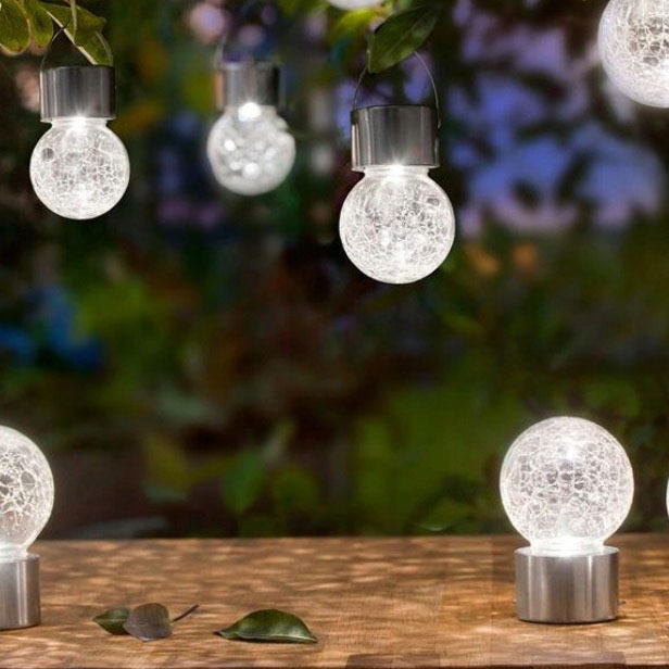 Lampka wisząca solarna Glovi 6000K, zimna biała, z przezroczystą kulą z efektem crackle. Produkt zawieszony na metalowych uchwytach, umieszczony w ogrodzie, z widocznymi detalami oświetlenia w tle.