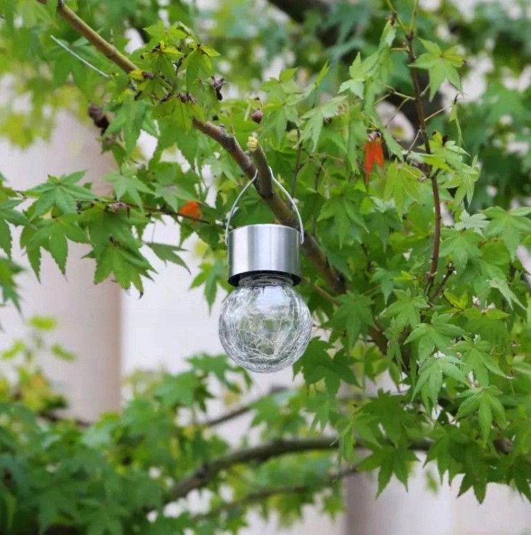 Lampka wisząca solarna Glovi 6000K w kształcie okrągłej, przezroczystej kulki, zawieszona na gałęzi drzewa. Posiada metalowy uchwyt oraz srebrną podstawę. Otoczona zielonymi liśćmi, co podkreśla jej naturalny kontekst użytkowania.