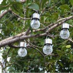 Lampka wisząca solarna Glovi 6000K w kolorze zimnej białej, z przezroczystym, teksturowanym kloszem i metalowym uchwytem. Cztery lampki zawieszone na gałęziach drzewa, w otoczeniu zieleni. Klosze mają okrągły kształt.