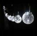 Lampka wisząca solarna z kulistym kloszem, wykonana z przezroczystego, teksturowanego szkła, emitująca zimne białe światło (6000K). Widoczne kilka kul zawieszonych w linii na ciemnym tle, z efektem pękniętej powierzchni.
