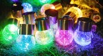 Lampka wisząca solarna RGB z przezroczystą kulą, emitująca różnokolorowe światło (zielone, niebieskie, różowe, żółte) z metalowym, srebrnym uchwytem. Ułożone na trawie, w otoczeniu dekoracyjnych kwiatów.