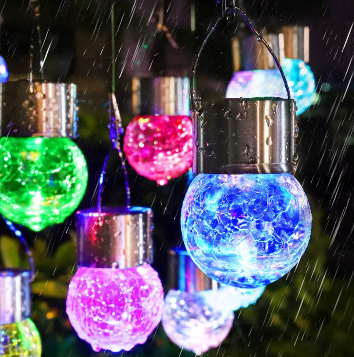 Lampka wisząca solarna RGB w kształcie szklanej kuli z efektem pękniętej struktury, świecąca w różnych kolorach (zielony, niebieski, różowy, fioletowy). Wisi na metalowym uchwycie, w otoczeniu deszczu.