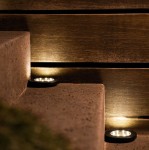 Czarna lampa solarna ogrodowa Terra wbijana, widoczna na schodach, emitująca delikatne światło. Szeroka podstawa i okrągły kształt, umieszczona na tle drewnianej ściany. Idealna do oświetlenia ogrodu lub ścieżek.