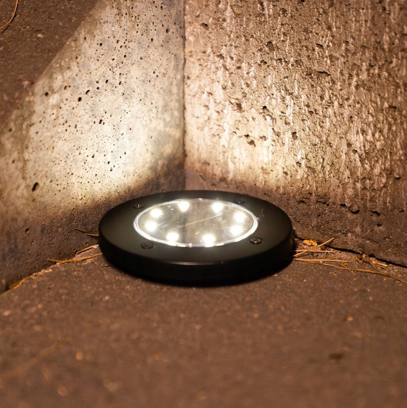 Lampa solarna Terra wbijana, czarna, okrągła, z sześcioma diodami LED. Umieszczona w narożniku, na ciemnej powierzchni, emitująca delikatne światło. Widoczne elementy montażowe i tekstura obudowy.