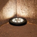 Lampa solarna Terra wbijana, czarna, okrągła, z sześcioma diodami LED. Umieszczona w narożniku, na ciemnej powierzchni, emitująca delikatne światło. Widoczne elementy montażowe i tekstura obudowy.
