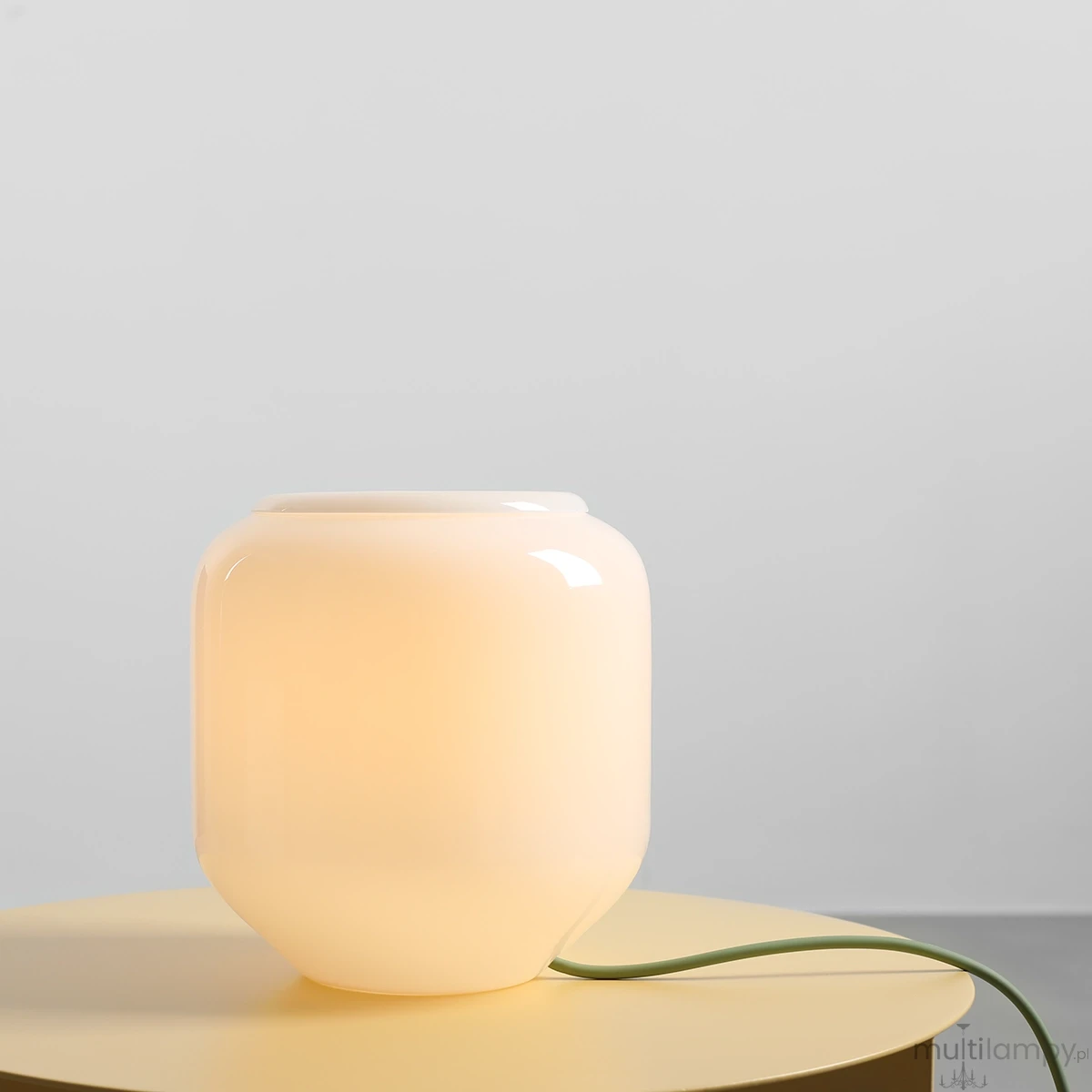 Lampa stołowa Flask Sage 1xE14 w kolorze białym i szałwiowym, o zaokrąglonym kształcie. Wykonana z przezroczystego materiału, emituje ciepłe światło. Ułożona na okrągłym stole, z widocznym zielonym kablem. Tło jednolite, szare.