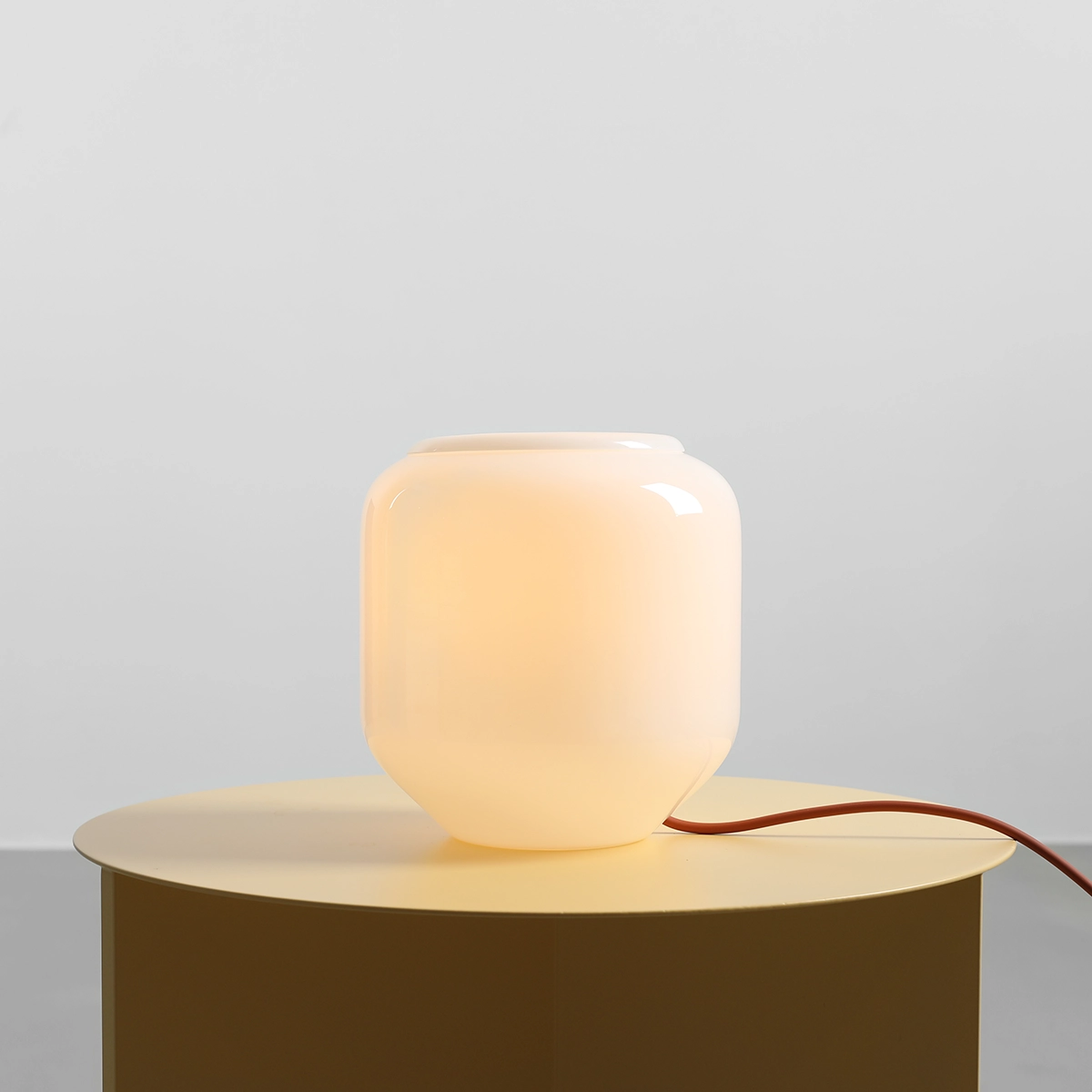 Lampa stołowa Flask Terra, o kształcie zaokrąglonego słoika, w kolorze białym z ceglanym przewodem. Ustawiona na okrągłym, złotym stoliku, emituje ciepłe światło, tworząc przytulną atmosferę.