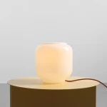 Lampa stołowa Flask Terra, o kształcie zaokrąglonego słoika, w kolorze białym z ceglanym przewodem. Ustawiona na okrągłym, złotym stoliku, emituje ciepłe światło, tworząc przytulną atmosferę.