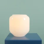 Lampa stołowa Flask Sapphire w kształcie zaokrąglonego pojemnika, wykonana z białego, błyszczącego szkła. Ustawiona na niebieskim stoliku, z widocznym górnym otworem. Idealna do oświetlenia wnętrz, z miejscem na żarówkę E14.