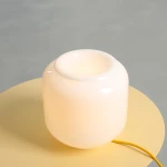 Lampa stołowa Flask Mimosa w kolorze białym z żółtym kablem, o zaokrąglonym, szklanym kształcie. Ujęcie z góry, widać otwór na żarówkę E14, lampa umieszczona na okrągłym, żółtym blacie.