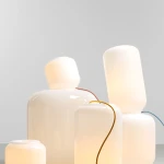 Lampa stołowa Flask Mimosa w białym kolorze z żółtym przewodem. Owalne i cylindryczne kształty, szklana konstrukcja, emitująca miękkie światło. Ułożona w grupie, podkreślająca nowoczesny design.