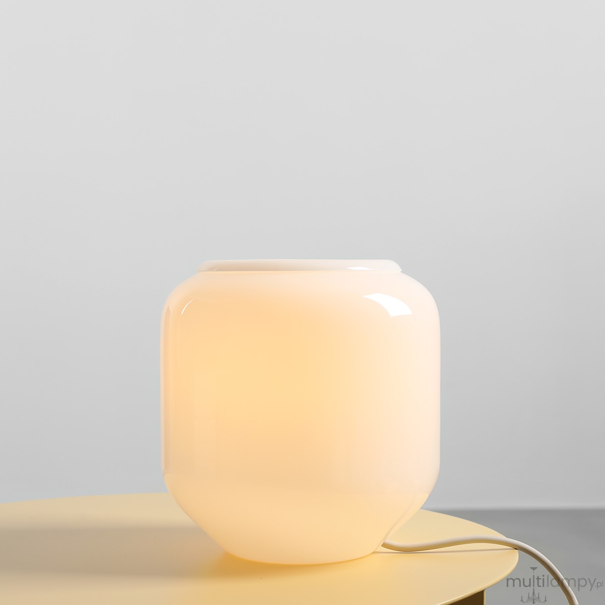 Lampa stołowa Flask Cotton w kolorze białym i kremowym, wykonana z gładkiego materiału. Ma zaokrąglony kształt z delikatnym podświetleniem. Ujęcie z boku, stojąca na okrągłym stole. Widoczny kabel zasilający. Tło jasne, minimalistyczne.
