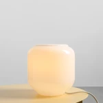 Lampa stołowa Flask Cotton w kolorze białym i kremowym, wykonana z gładkiego materiału. Ma zaokrąglony kształt z delikatnym podświetleniem. Ujęcie z boku, stojąca na okrągłym stole. Widoczny kabel zasilający. Tło jasne, minimalistyczne.