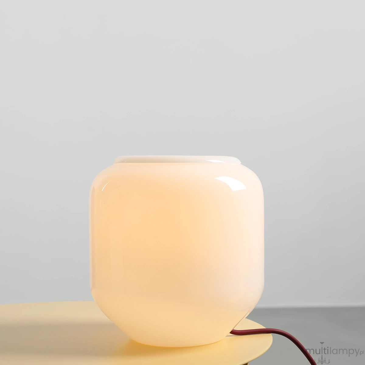 Lampa stołowa Flask Cherry z kloszem w kolorze białym, o zaokrąglonym kształcie, wykonana z transparentnego szkła. Widoczny czerwony kabel zasilający. Ujęcie z boku na tle szaro-białym. Przeznaczona na żarówkę E14.