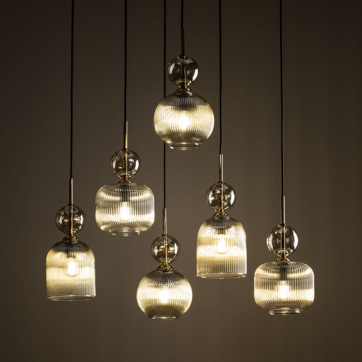 Lampa wisząca Sophia z sześcioma kloszami E14 w kolorze bursztynowym. Klosze mają różne kształty, są szklane z żłobieniami, zawieszone na czarnych linkach. Błyszczące detale metalowe podkreślają elegancję lampy. Tło w odcieniu ciemnym.