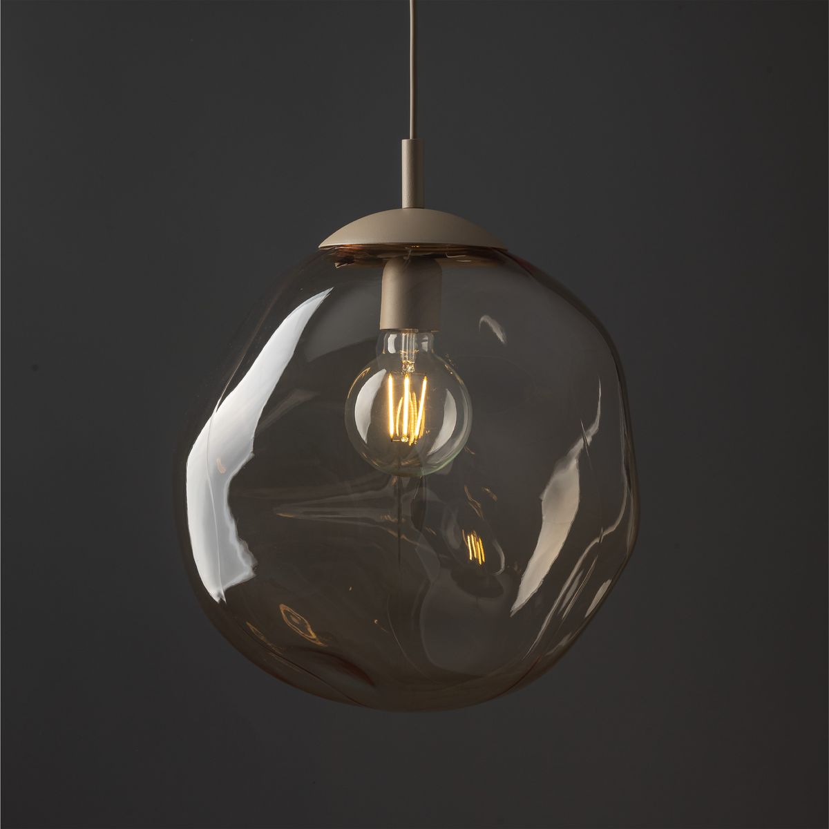 Lava Sabia D35 lampa wisząca w kolorze beżowym z przezroczystym, organicznie uformowanym kloszem. Widoczna żarówka E27, zawieszona na cienkim przewodzie. Tło ciemne, co podkreśla elegancki design lampy.
