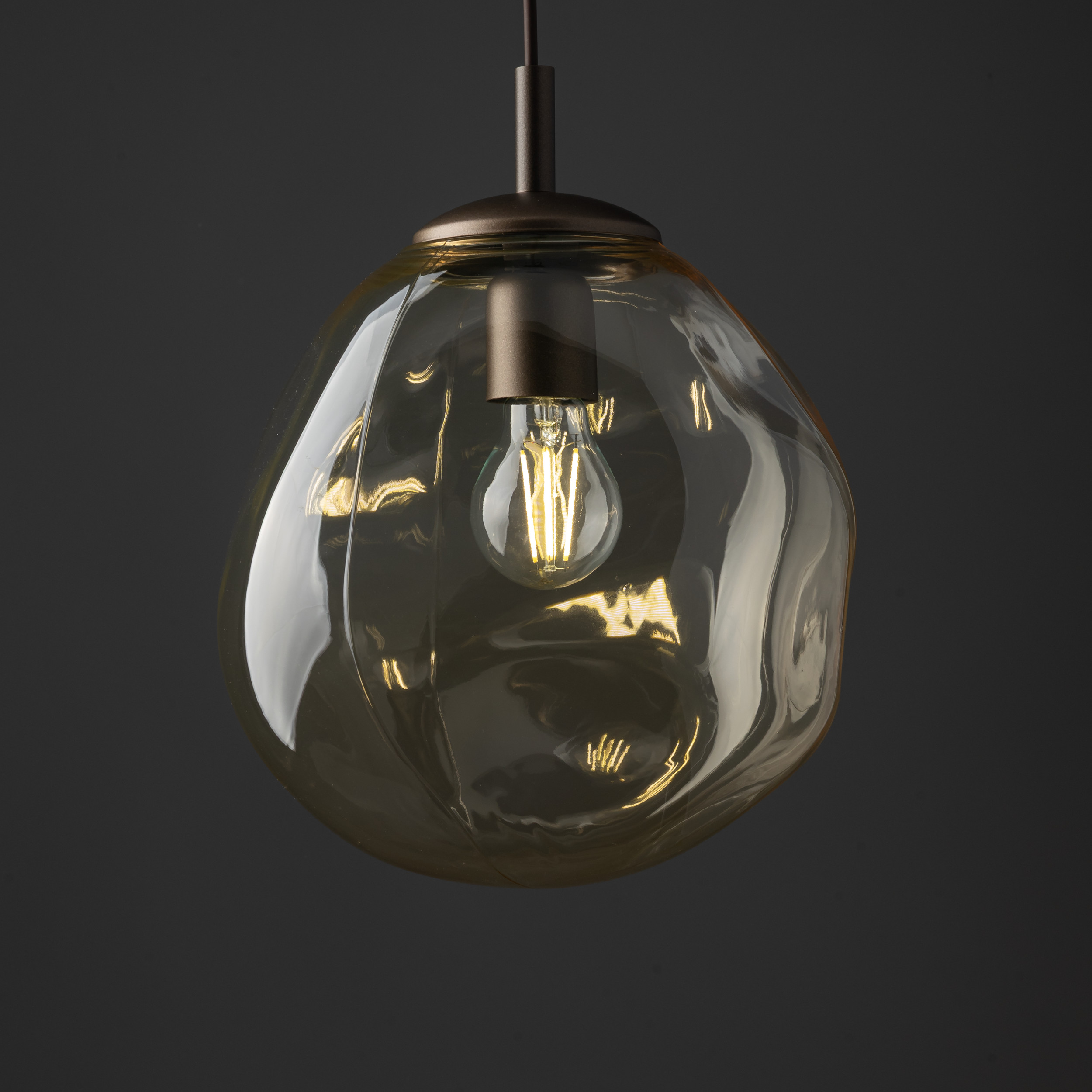 Lava Brown D25 to brązowa lampa wisząca z kloszem w kształcie kropli, wykonana z przezroczystego szkła. W centralnej części znajduje się żarówka E27. Lampa zawieszona na stylowym, metalowym mocowaniu w ciemnym kolorze, na tle szarym.
