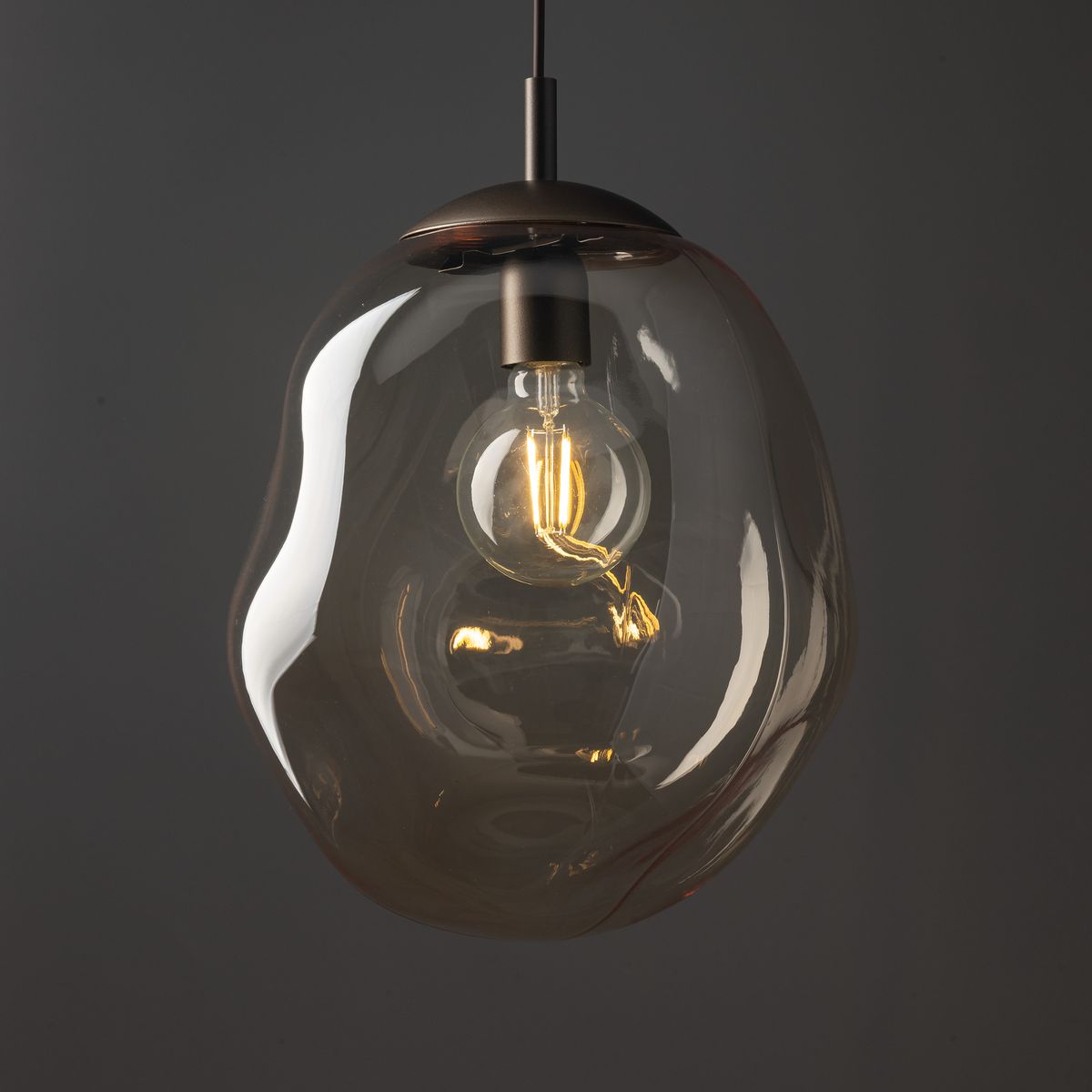 Brązowa lampa wisząca Lava Brown D35, z przezroczystym kloszem o organicznym kształcie, zawieszona na cienkim przewodzie. Wewnątrz znajduje się żarówka E27, emitująca ciepłe światło. Tło ciemne, podkreślające elegancję produktu.