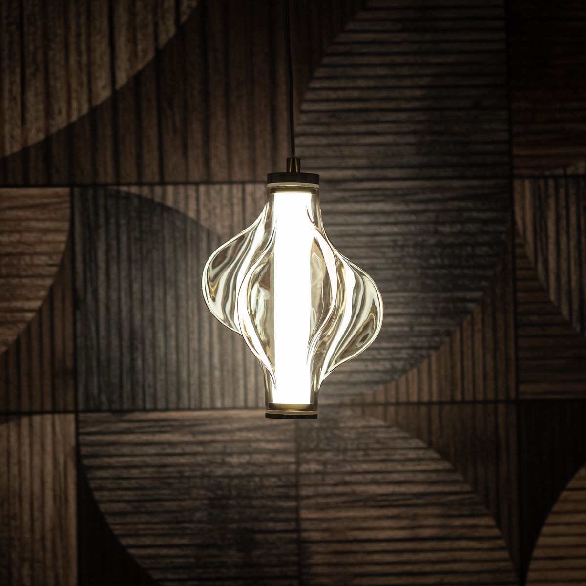 Lampa wisząca LED Elva 8W 3000K w kolorze mosiądzu z bursztynowym kloszem, o organicznym kształcie z falującymi liniami, zawieszona na cienkim przewodzie, na tle drewnianej ściany z geometrycznym wzorem.