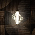 Lampa wisząca LED Elva 8W 3000K w kolorze mosiądzu z bursztynowym kloszem, o organicznym kształcie z falującymi liniami, zawieszona na cienkim przewodzie, na tle drewnianej ściany z geometrycznym wzorem.
