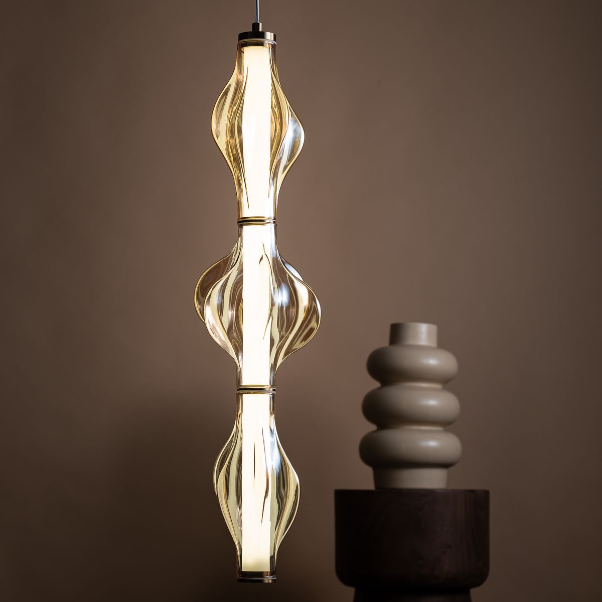 Lampa wisząca LED Elva 22W 3000K w kolorze mosiądzu i bursztynu, o unikalnym, falistym kształcie. Zawieszona w pionie, emituje ciepłe światło. W tle widoczny ceramiczny wazon w kształcie spirali na drewnianym stoliku.