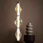Lampa wisząca LED Elva 22W 3000K w kolorze mosiądzu i bursztynu, o unikalnym, falistym kształcie. Zawieszona w pionie, emituje ciepłe światło. W tle widoczny ceramiczny wazon w kształcie spirali na drewnianym stoliku.