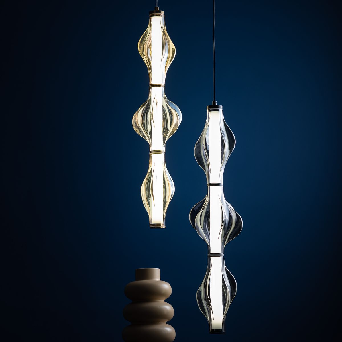 Lampa wisząca LED Elva 22W 3000K w kolorze mosiężnym i bursztynowym, z dwoma szklanymi kloszami o falistym kształcie, zawieszona na cienkim przewodzie, w tle ciemnoniebieskiej ściany. W dolnej części widoczny ceramiczny element dekoracyjny.