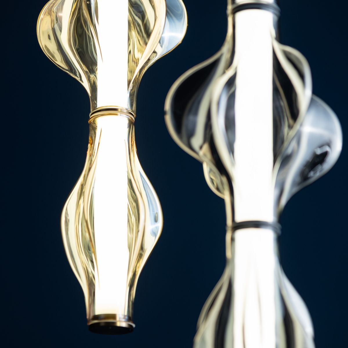Lampa wisząca LED Elva o mocy 22W, kolor mosiężny i bursztynowy. Zawiera faliste, przezroczyste klosze z wbudowanym źródłem światła, zawieszona na cienkim przewodzie. Tło ciemnoniebieskie, podkreślające elegancki design lampy.