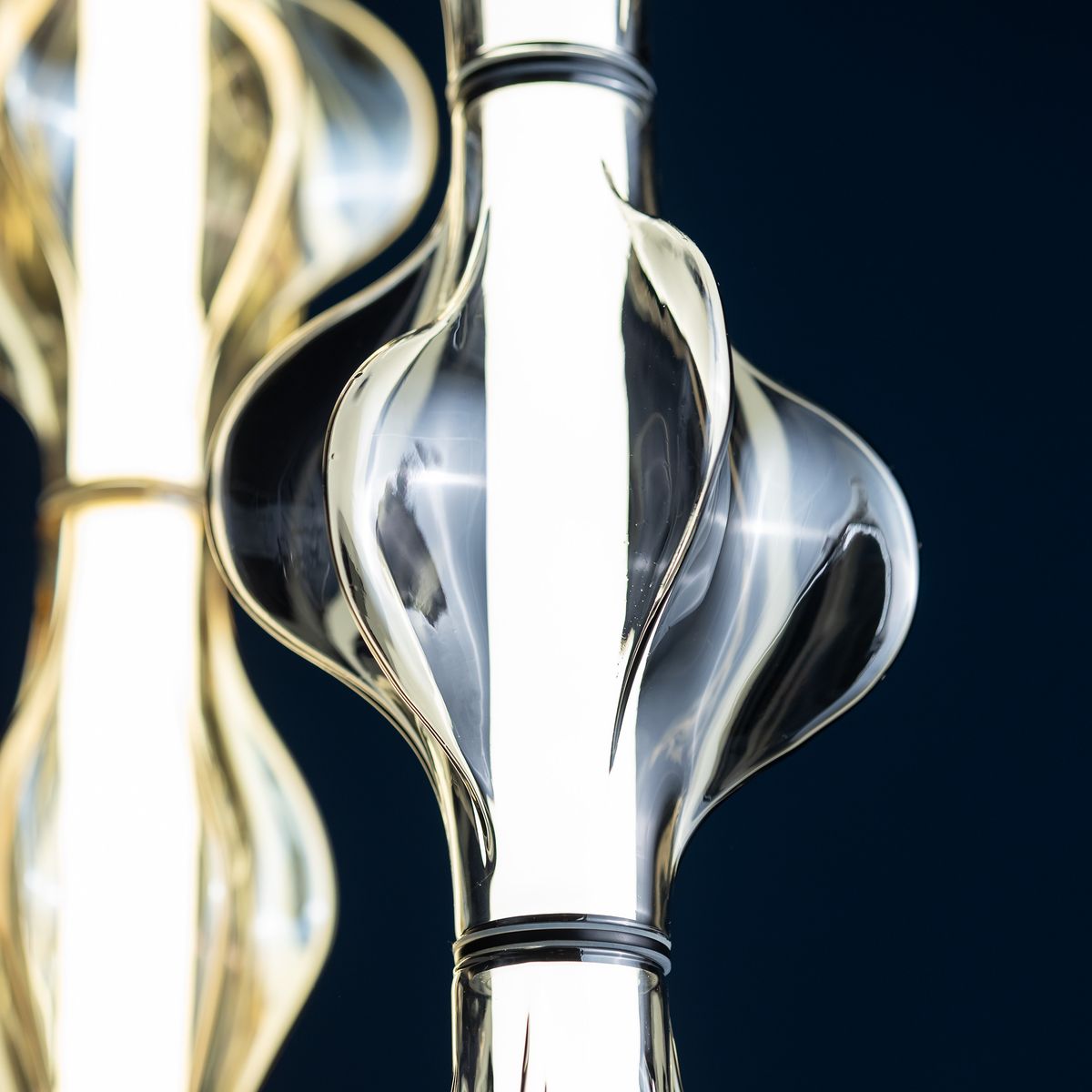 Lampa wisząca Elva LED 22W 3000K w kolorze mosiądzu i bursztynu, z efektownym, falistym kloszem ze szkła, umieszczona w centralnej części, z widocznym źródłem światła w formie długiej listwy. Tło ciemnoniebieskie.