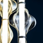 Lampa wisząca Elva LED 22W 3000K w kolorze mosiądzu i bursztynu, z efektownym, falistym kloszem ze szkła, umieszczona w centralnej części, z widocznym źródłem światła w formie długiej listwy. Tło ciemnoniebieskie.