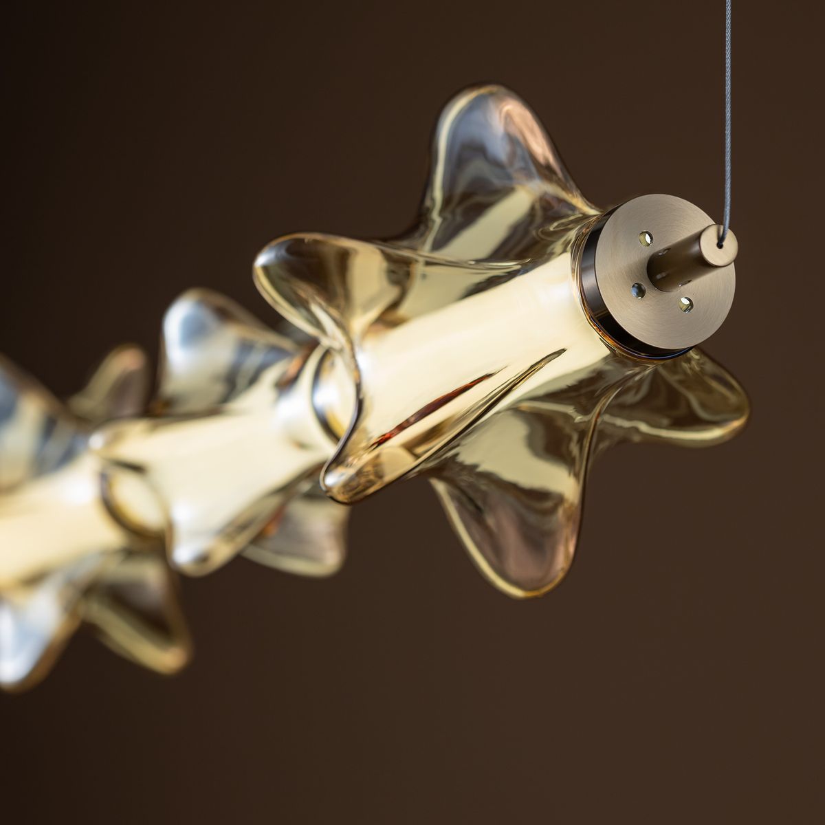 Lampa wisząca LED Elva 30W z bursztynowym kloszem w kształcie gwiazd, z metalowym mocowaniem w kolorze mosiądzu. Zbliżenie na detal klosza z widocznym światłem LED, zawieszona na cienkim przewodzie. Tło w ciemnym odcieniu.