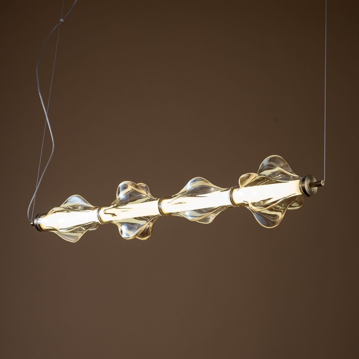 Lampa wisząca LED Elva 30W, 3000K, w kolorze mosiądzu i bursztynu. Wyposażona w szklane klosze o falistym kształcie, zawieszona na cienkim przewodzie, z widocznym źródłem światła w centralnej części. Tło w stonowanym odcieniu.